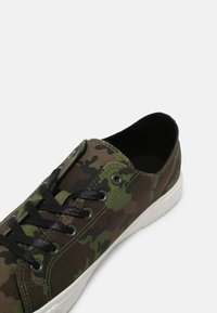 Baskets basses avec motif camouflage vert et marron, lacets noirs, œillets métalliques et semelle en caoutchouc blanche.