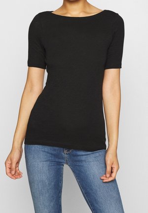 Mujer con una camiseta negra ajustada de manga corta y vaqueros azules, con un fondo gris neutro.