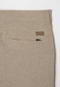 Pantalons en tissu beige avec une finition texturée, dotés d'une poche arrière avec une fermeture éclair et d'une petite étiquette de marque marron cousue au-dessus.