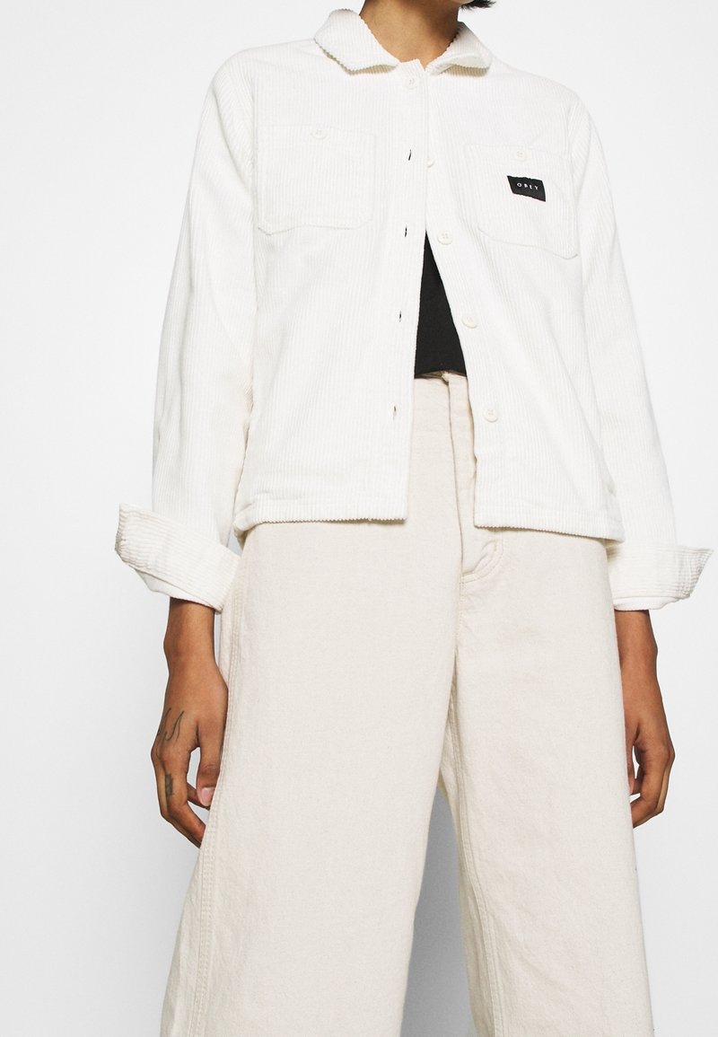 Vit kordjacka med krage, två framfickor och en svart logotyp-patch, tillsammans med krämfärgade vida byxor.