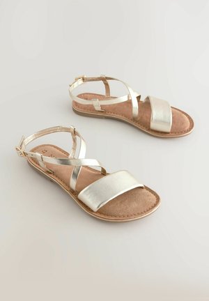 Fém arany szandál, lapos talppal, széles elülső pánttal és keresztbe futó bokapántokkal, érdes, barnás lábfejre helyezve.