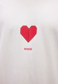 Λευκό ύφασμα με κόκκινο γεωμετρικό σχέδιο καρδιάς πάνω α πό τη λέξη "HUGO" τυπωμένη με κόκκινο, στο κέντρο της περιοχής του στήθους.