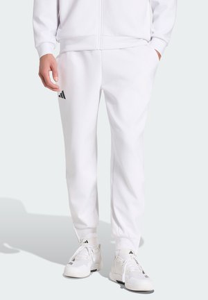 Personne portant un pantalon de survêtement Adidas blanc, des chaussures de sport Adidas blanches et une veste blanche zippée, debout avec une main dans la poche.