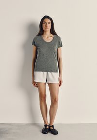 Moteris stovi priešais paprastą šviesią sieną, vilkinti pilką marškinėlį, šviesiai pilkus šortus ir juodus sandalus su uždarytais pirštais, žvelgia tiesiai į priekį su neutraliu veido išraiška.