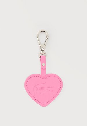 Porte-clés en cuir rose en forme de cœur avec logo crocodile embossé et fermoir en métal argenté sur fond clair.