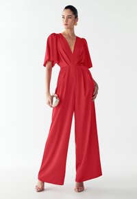 Red jumpsuit with a deep V-neckline, short flutter sleeves, and wide-leg trousers. Smooth fabric with a lightweight texture. Gold clutch in hand.