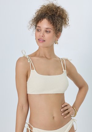 Frau mit lockigem Haar in einem cremefarbenen, texturierten Bikinioberteil und -unterteil, die seitlich blickt und die Hand in der Hüfte hat, vor einem schlichten hellen Hintergrund.