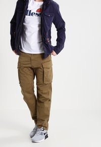 Giacca navale con tasche automatiche, t-shirt bianca con logo, pantaloni cargo oliva con più tasche e sneakers grigie Nike.