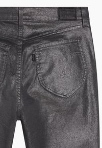Jean gris métallisé avec une texture légèrement brillante. Présente un design classique à cinq poches et une étiquette de marque en cuir sur la ceinture arrière.