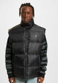 Sort puffervest med høj krave, frontlynlås og quiltet design. Har et lille logo og bæres over en stribet sweater.