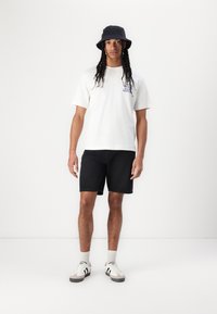 Tricou alb din bumbac cu grafică pe față, pantaloni scurți negri din denim, pălărie de tip bucket neagră și adidași albi cu accente negre, stând pe un fundal neutrals.