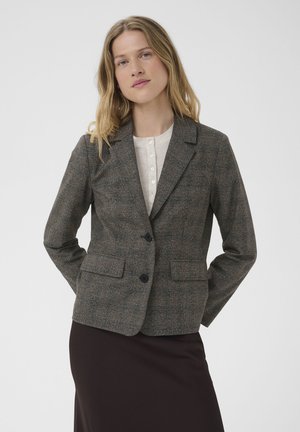Femme portant un blazer gris à carreaux sur une blouse blanche et une jupe foncée, debout les mains dans le dos devant un fond uni.