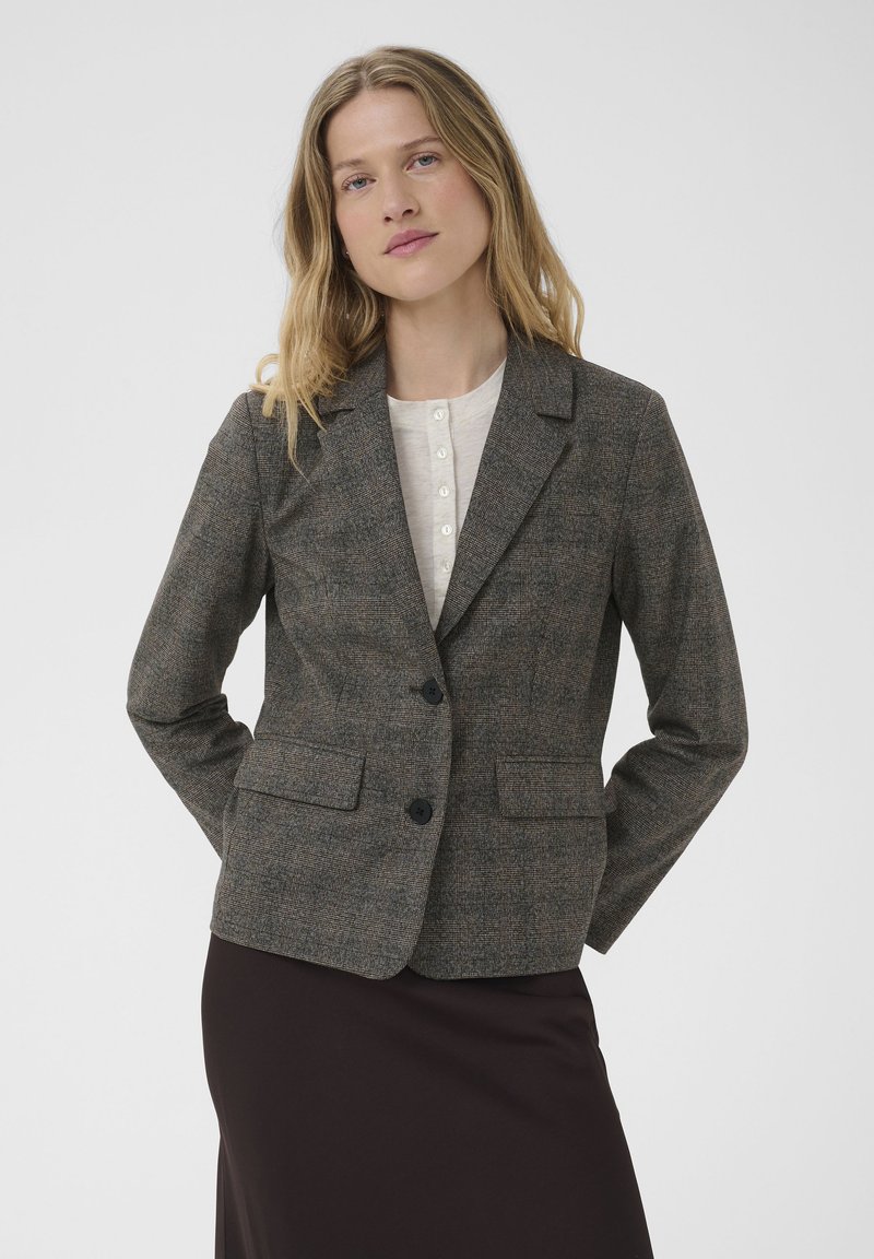 Femme portant un blazer gris à carreaux sur une blouse blanche et une jupe foncée, debout les mains dans le dos devant un fond uni.