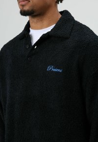 Schwarzer Fleecepullover mit strukturierter Oberfläche, ausgestattet mit einem Kragen, zwei Knöpfen und dem in Blau gestickten "Present"-Logo auf der Brust.