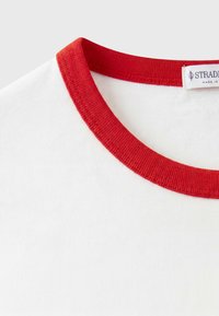 T-shirt blanc avec un col rond côtelé rouge et une étiquette de marque visible à l'intérieur du col.