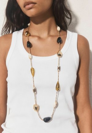 Woman wearing white sleeveless top with long necklace featuring black, gold, amber, and white irregular beads on a gold chain.