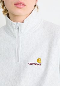Grå fleecegenser med høy krage, med glidelås foran og brodert gult og burgunder Carhartt-logo på brystet.