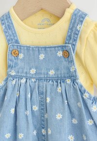 Żółta teksturowana koszulka dla niemowląt pod niebieską sukienką dżinsową z nadrukiem białych stokrotek i brązowymi guzikami na drewnianym wieszaku.