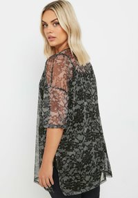Top in pizzo nero con maniche trasparenti, motivo floreale, vestibilità ampia e spacchi laterali. Tessuto testurizzato con un aspetto delicato e semitrasparente.