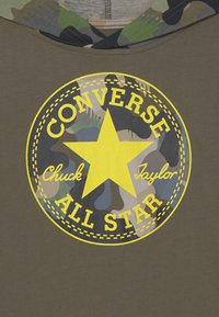 Zielona bluza z kapturem z wyściółką w kamuflażowy wzór, z dużą żółtą gwiazdą i okrągłym logo, które brzmi "Converse Chuck Taylor All Star."
