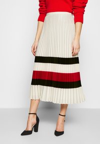 Falda midi plisada en crema con rayas horizontales rojas, negras y blancas. Tejido suave con una textura lisa, combinada con zapatos de tacón negro.