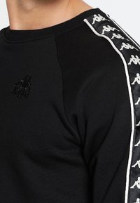 Svart sweatshirt med broderad logotyp, kontrasterande svarta axelpaneler och vitt Kappa-logotape längs ärmarna. Mjuk textur.