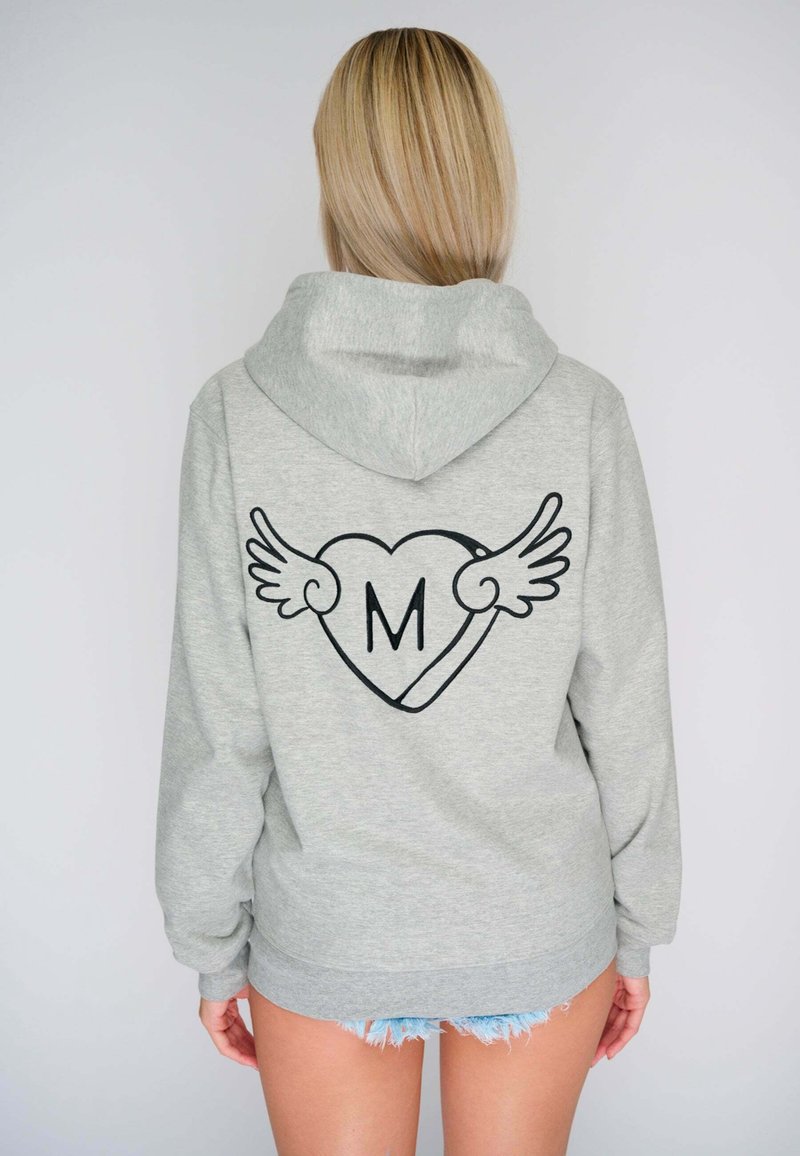 Personne blonde portant un sweat à capuche gris clair avec un cœur ailé noir et la lettre « M » au dos, assorti à un short en jean effiloché.