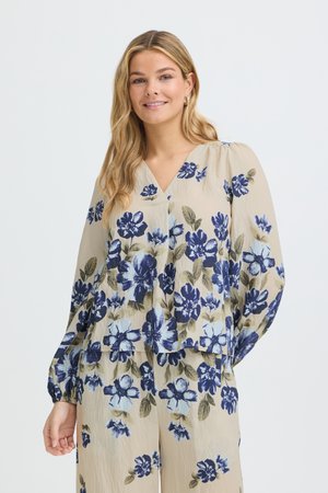 Kvinde med langt blondt hår iført beige bluse og bukser med blå og grønne blomsterprint, der smiler mod en ensfarvet hvid baggrund.