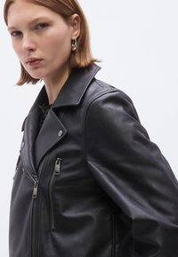 Schwarze Leder-Motorradjacke mit weitem Kragen, Reißverschlusstaschen und silbernen Beschlägen. Glatte Textur mit strukturierter Passform.