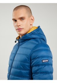 Kék szemű férfi, aki kék, steppelt kapucnis dzsekit visel mustársárga béléssel és "Big Star" felvarróval az ujjon, a kamerába néz.