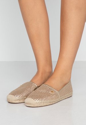 Stopy w noszonych złotych slip-onach espadrylach z metalicznym splotem, z podeszwami owiniętymi jutą, na jednolitym tle.