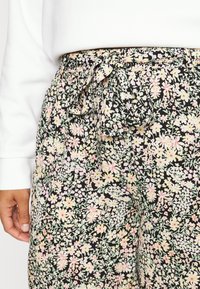 Pantaloni in tessuto nero con stampa floreale presentano una vita aderente con un fiocco legato. Il design include piccoli fiori rosa e gialli su steli verdi.