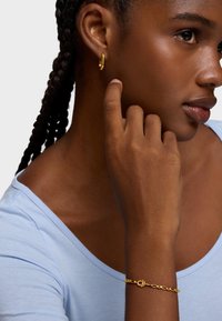 Złoty kolczyk-koło i łańcuszkowa bransoletka na ciemnosk�órej dłoni. Bransoletka ma zapięcie. Jasnoniebieski top z dekoltem w kształcie litery U.