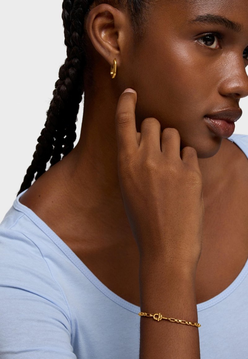 Złoty kolczyk-koło i łańcuszkowa bransoletka na ciemnoskórej dłoni. Bransoletka ma zapięcie. Jasnoniebieski top z dekoltem w kształcie litery U.