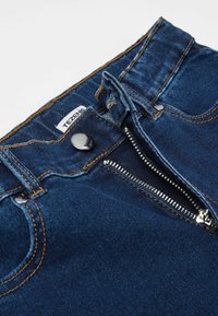 Tmavě modré džíny z denimu s mosazným knoflíkem, zapínáním na zip a hnědými švy. Na štítku je nápis "TEZENIS." Detailní pohled na pas.