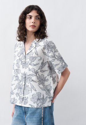 Γυναίκα με σγουρά μαλλιά που φοράει λευκό κοντομάνικο πουκάμισο με κουμπιά και μαύρα λουλουδάτα μοτίβα, καθώς και μπλε τζιν με λεπτομέρειες από πέρλες.