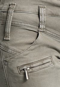 Olivenfarvede denim jeans med en frontlomme med lynlås, synlige sømme og bæltestropper, der fremhæver en blød tekstur.