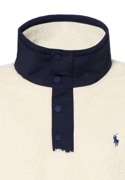 High collar cream fleece pullover with navy snap buttons and navy inner collar, featuring a small embroidered logo on the chest.