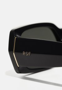 Mustad mustaga raamiga päikeseprillid julge ja nurkadega disainiga, mille templil on kuldne "RSF" logo ja tumedad sillerdavad läätsed.