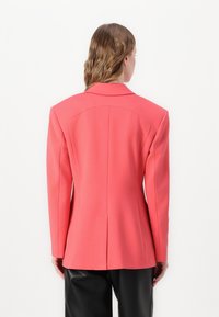 Koraalkleurige blazer met een gestructureerd ontwerp, enkele rugventilatie en scherpe revers. Gemaakt van een soepele stof, gecombineerd met zwarte broeken.