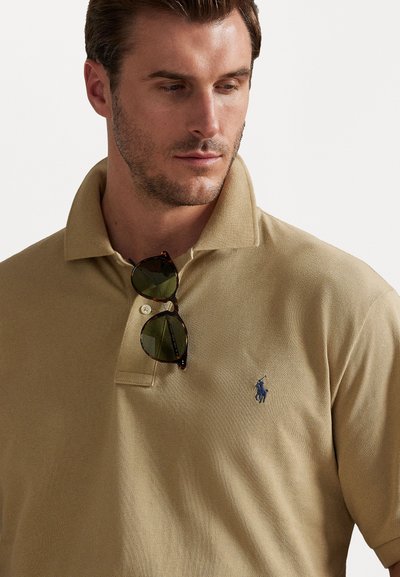 Polo shirt bege com uma superfície texturizada, mangas curtas e um logotipo azul. Óculos de sol repousam na gola, com uma armação em padrão tartaruga.