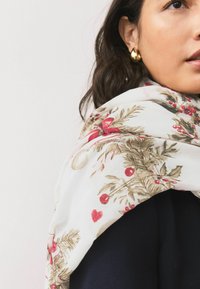 Lehký bílý šátek s veselým vzorem červených bobulí, zelených listů a zlatých ozdob, přehodil přes tmavý top.