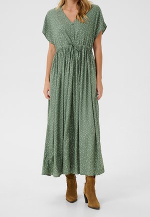Femme portant une robe verte à manches courtes, boutonnée, longue jusqu'aux chevilles, avec des pois blancs et des bottes à talons en daim marron.