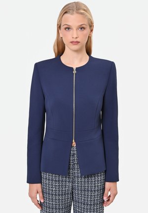 Giovane donna con lunghi capelli biondi che indossa una giacca blu navy aderente con cerniera frontale e pantaloni a fantasia bianco e nero.
