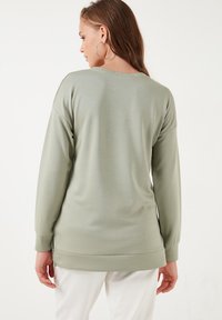 Top de manga larga verde claro con un corte relajado, escote redondeado y aberturas laterales en el dobladillo. Textura suave con un brillo sutil.
