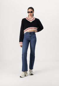 Svart fluffig kort tröja med röd och vit randig kant, ihop med högmidjade blå jeans och beigea sportskor. Mörka solglasögon på.
