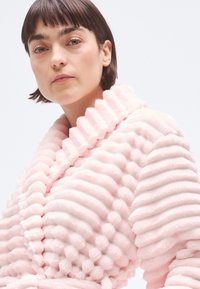 Halat de pluș roz cu textură nervurată și guler cu volane. Țesătura pare moale și groasă, având o culoare uniformă și netedă pe toată suprafața.