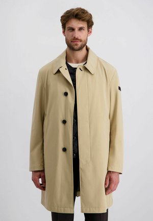 Mann mit braunen Haaren und Bart, der einen beigen einreihigen Mantel über einer schwarzen gesteppte Jacke trägt, steht vor einem schlichten grauen Hintergrund.