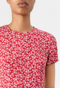 Chemise à motifs floraux rouges avec des manches courtes, un col rond et un petit logo en détail. Présente des fleurs blanches et des accents bleu clair.
