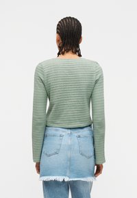 Άτομο με πλεγμένα μαλλιά που φοράει μπλούζα μακριών μανικιών σε υφή χλωμού πράσινου και φθαρμένη φούστα από τζιν ανοιχτού μπλε χρώματος πάνω από μπλε τζιν, με την πλάτη προς τα εμάς.